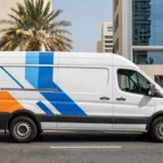 A white commercial van with professional blue and orange abstract branding parked on a sunlit street in Dubai with modern buildings in the background.